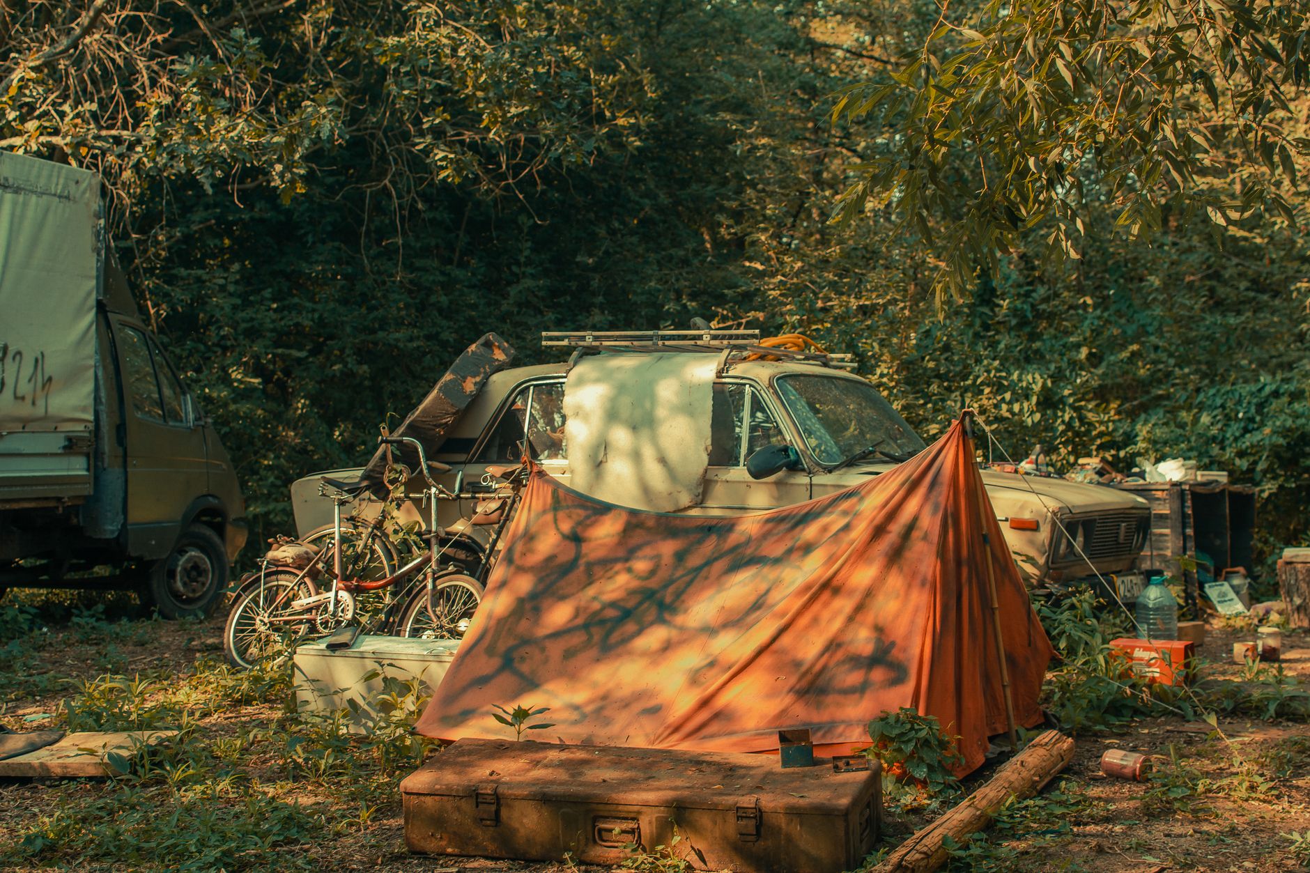 A vintage car in a rustic campsite surrounded by trees and an orange tent.